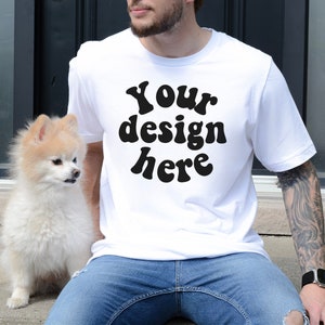 Può includere: Una maglietta bianca con il testo "Your design here" in lettere nere in stile retrò. La maglietta è indossata da una persona con tatuaggi sul braccio. Un piccolo cane bianco è seduto accanto alla persona.