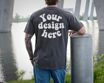 May include: Dark grey t-shirt with the text "Your design here" in white, curved across the back. The shirt is worn with blue jeans. The person is leaning on a concrete post near water.