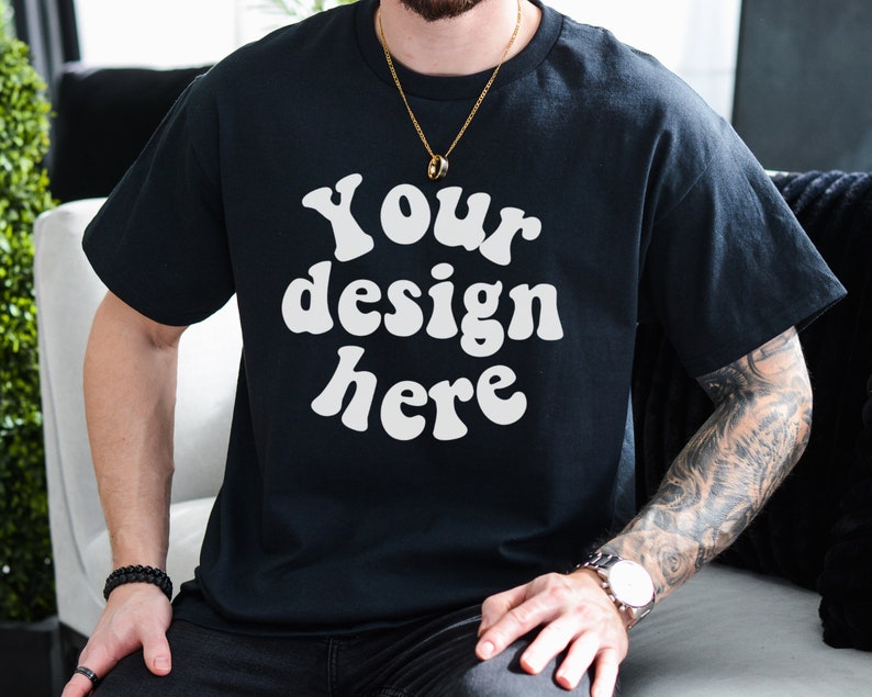 May include: A black t-shirt with the text "Your design here" in white, retro-style lettering. The shirt is being worn by a person with a gold chain necklace and a tattoo on their arm.