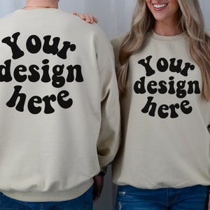 May include: Two cream-coloured sweatshirts with the text "Your design here" in black, retro-style font. The sweatshirts are shown on two people, one standing behind the other.