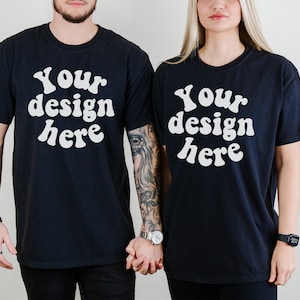 K&ouml;nnte beinhalten: Zwei schwarze T-Shirts mit dem Text "Your design here" in wei&szlig;er, retro-artiger Schrift. Die Shirts werden von zwei Personen getragen, die nebeneinander stehen.