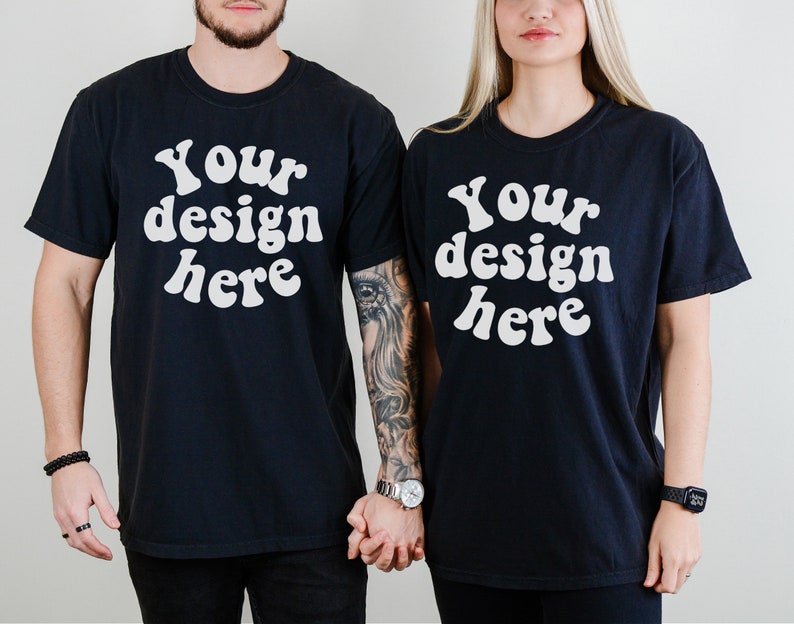 K&ouml;nnte beinhalten: Zwei schwarze T-Shirts mit dem Text "Your design here" in wei&szlig;er, retro-artiger Schrift. Die Shirts werden von zwei Personen getragen, die nebeneinander stehen.
