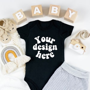 May include: Black baby onesie with the text "Your design here" in white. The onesie is surrounded by wooden blocks spelling "BABY", a rainbow-colored yarn wall hanging, a pair of baby shoes, and a toy camera.
