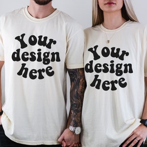 May include: Two cream-colored t-shirts with the text "Your design here" in a black, retro font. The shirts are short-sleeved and appear to be made of a soft material. The image shows two people wearing the shirts.