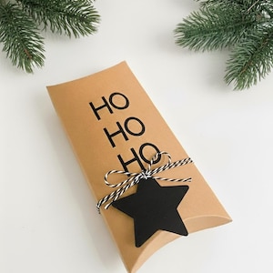 Könnte beinhalten: Eine rechteckige Geschenkbox aus Kraftpapier mit den Wörtern "HO HO HO" in Schwarz aufgedruckt. Die Box ist mit schwarz-weißem Bindfaden und einem schwarzen sternförmigen Anhänger gebunden. Grüne Tannenzweige sind im Hintergrund.