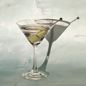 May include: A clear martini glass filled with a clear liquid and two green olives. A silver cocktail pick holds the olives. The glass casts a shadow on the light blue and white background. The drink is a classic cocktail.
