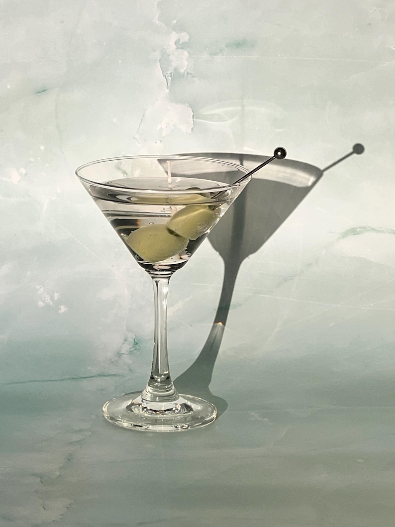 May include: A clear martini glass filled with a clear liquid and two green olives. A silver cocktail pick holds the olives. The glass casts a shadow on the light blue and white background. The drink is a classic cocktail.