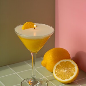 May include: A candle shaped like a cocktail in a martini glass, with a lit wick and a lemon slice garnish. The candle has a yellow base and a white top, resembling a drink. Two lemons, one halved, are next to the glass.