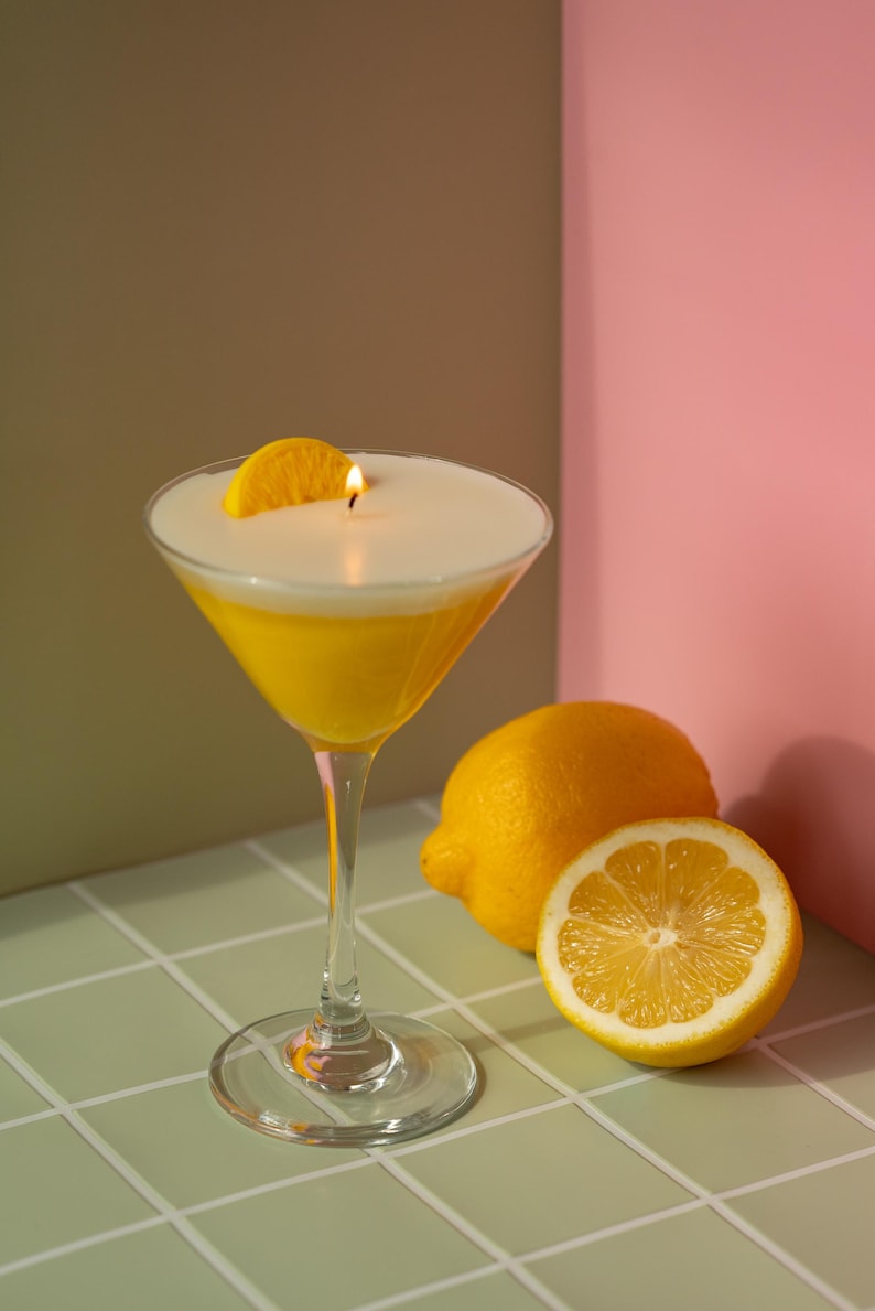 May include: A candle shaped like a cocktail in a martini glass, with a lit wick and a lemon slice garnish. The candle has a yellow base and a white top, resembling a drink. Two lemons, one halved, are next to the glass.