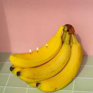 以下が含まれることがあります： 熟した黄色のバナナの束で、そのうちの1つには3本のろうそくが灯っています。バナナは、ピンク色の背景に対して、明るい緑色のタイルの表面にあります。ろうそくは、果物にユニークな装飾要素を追加します。
