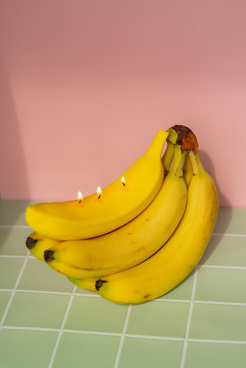 以下が含まれることがあります： 熟した黄色のバナナの束で、そのうちの1つには3本のろうそくが灯っています。バナナは、ピンク色の背景に対して、明るい緑色のタイルの表面にあります。ろうそくは、果物にユニークな装飾要素を追加します。
