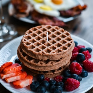 Puede incluir: Una pila de cuatro gofres de chocolate cubiertos con arándanos, frambuesas y fresas en rodajas en un plato blanco.
