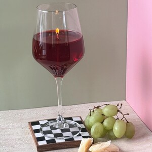 Peut inclure: Un grand verre &agrave; vin rempli de cire rouge, allum&eacute; avec une petite flamme, cr&eacute;ant une bougie. La bougie repose sur un dessous de verre &agrave; carreaux noir et blanc. Des raisins verts et du fromage sont dispos&eacute;s &agrave; proximit&eacute;.