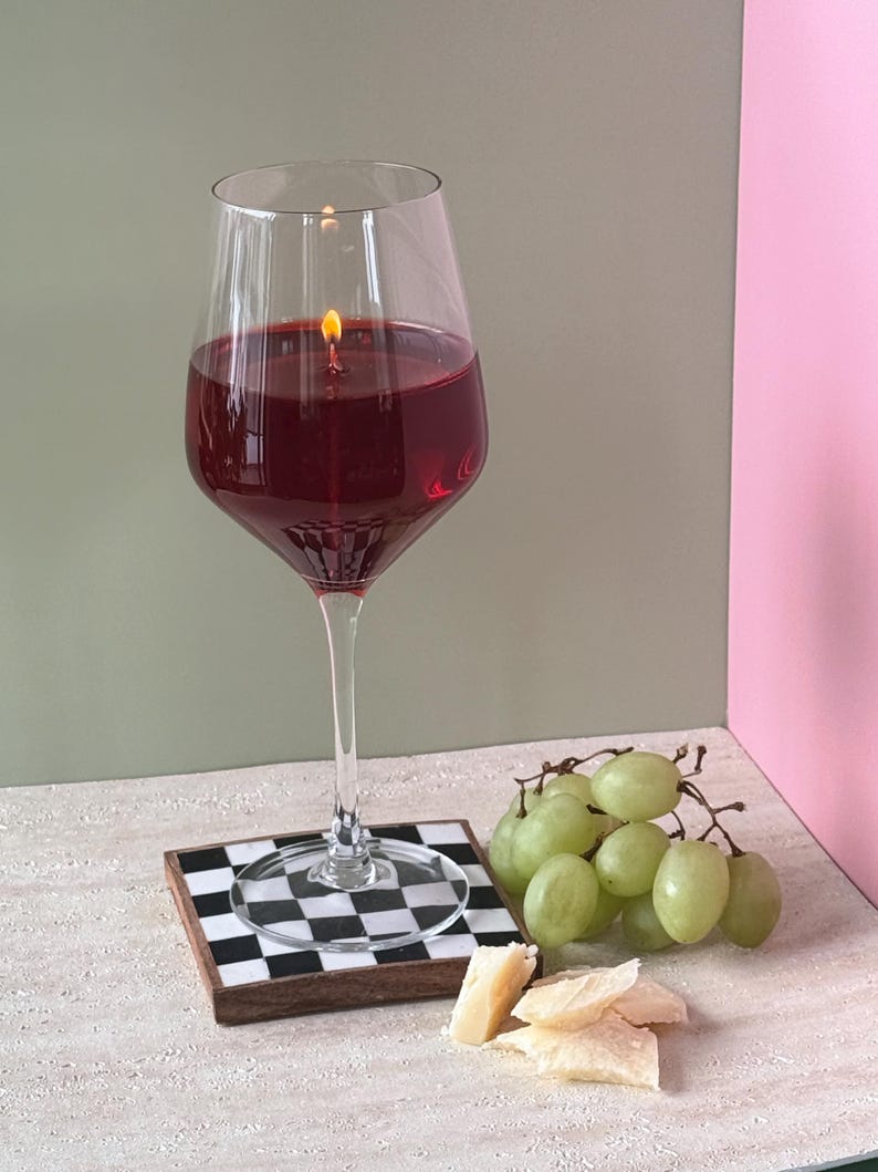 Peut inclure: Un grand verre &agrave; vin rempli de cire rouge, allum&eacute; avec une petite flamme, cr&eacute;ant une bougie. La bougie repose sur un dessous de verre &agrave; carreaux noir et blanc. Des raisins verts et du fromage sont dispos&eacute;s &agrave; proximit&eacute;.