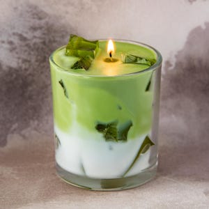May include: A candle in a clear glass, designed to resemble a matcha latte. The candle has a layered effect with white and green wax, and is topped with green wax pieces. A lit wick is visible.