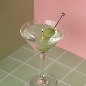 May include: A martini glass candle with a lit wick, featuring two green olives on a metal skewer. The candle is set against a backdrop of pink and olive green, with a tiled surface.