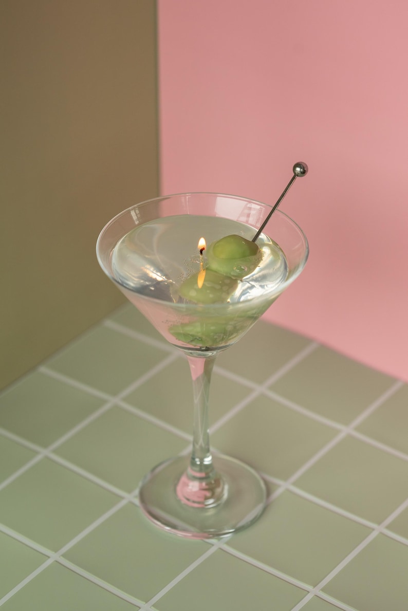 May include: A martini glass candle with a lit wick, featuring two green olives on a metal skewer. The candle is set against a backdrop of pink and olive green, with a tiled surface.