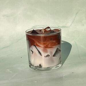 May include: A clear glass candle resembling an iced coffee. The candle has layers of white, brown, and dark brown wax, with wax cubes on top. A single wick is visible in the center. The candle sits on a light green surface.