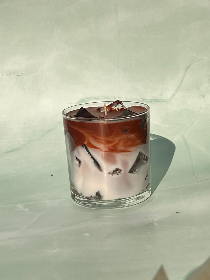 May include: A clear glass candle resembling an iced coffee. The candle has layers of white, brown, and dark brown wax, with wax cubes on top. A single wick is visible in the center. The candle sits on a light green surface.