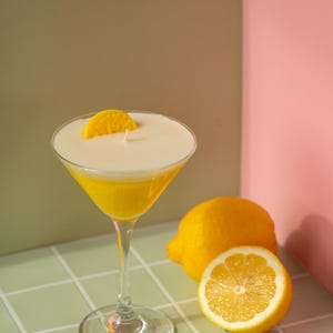 May include: A yellow cocktail candle in a martini glass, garnished with a lemon slice. A whole lemon and a lemon half are next to the glass. The background is a mix of green, brown, and pink.