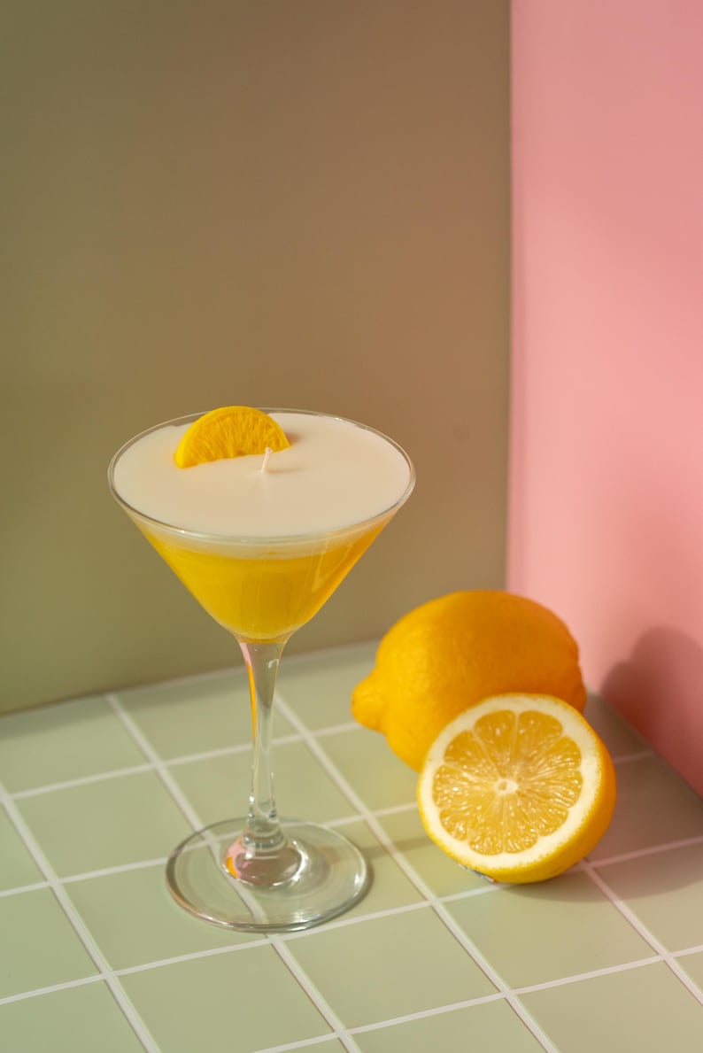May include: A yellow cocktail candle in a martini glass, garnished with a lemon slice. A whole lemon and a lemon half are next to the glass. The background is a mix of green, brown, and pink.