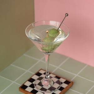 May include: A martini glass with a clear liquid, two green olives, and a metal cocktail pick. The glass sits on a checkered coaster with a wooden frame. The background is split between pink and olive green.