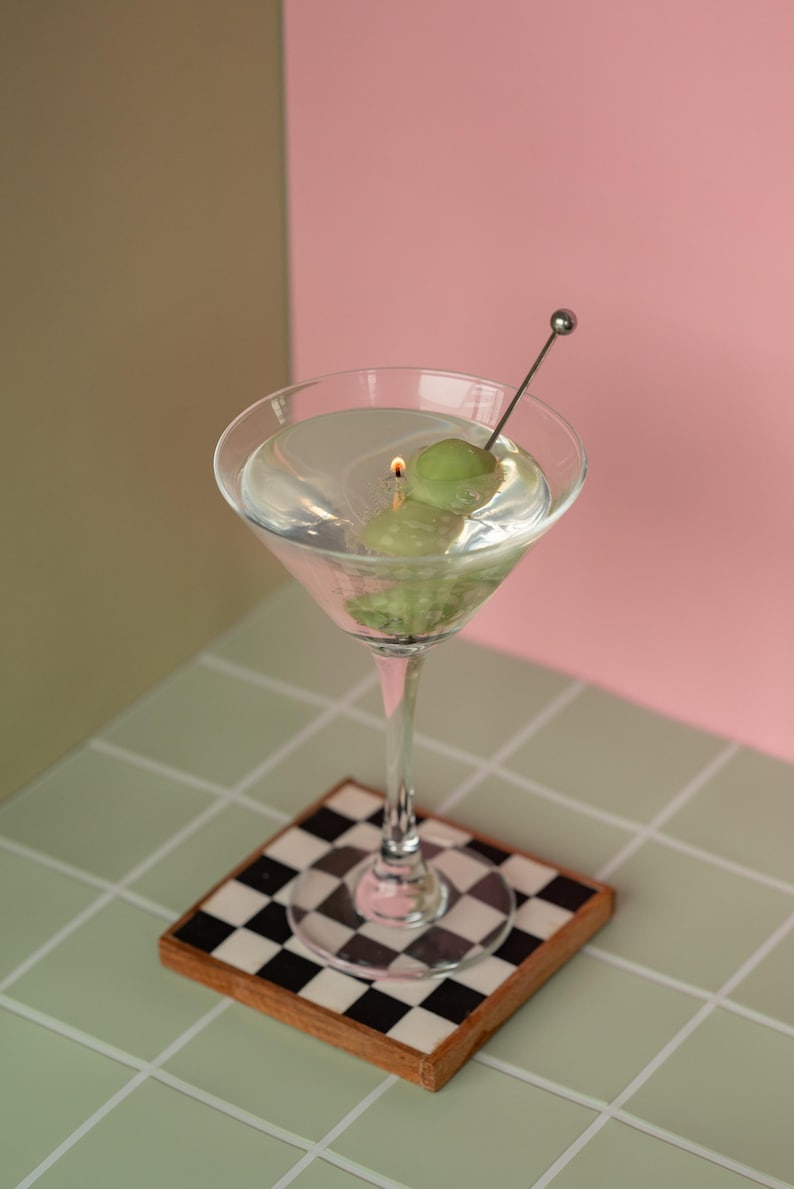 May include: A martini glass with a clear liquid, two green olives, and a metal cocktail pick. The glass sits on a checkered coaster with a wooden frame. The background is split between pink and olive green.