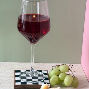 Peut inclure: Une bougie en forme de grand verre &agrave; vin remplie de cire rouge, avec une m&egrave;che allum&eacute;e. Le verre repose sur un dessous de verre &agrave; carreaux noir et blanc, &agrave; c&ocirc;t&eacute; d'un tas de raisins verts et de fromage.