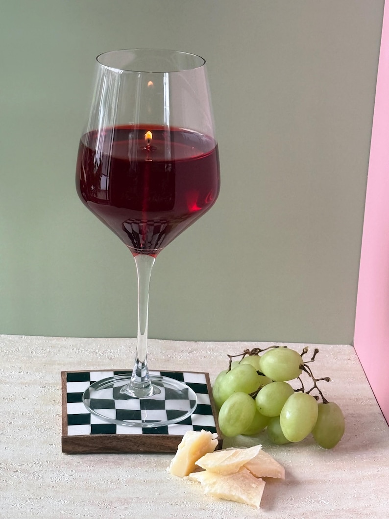 Peut inclure: Une bougie en forme de grand verre &agrave; vin remplie de cire rouge, avec une m&egrave;che allum&eacute;e. Le verre repose sur un dessous de verre &agrave; carreaux noir et blanc, &agrave; c&ocirc;t&eacute; d'un tas de raisins verts et de fromage.