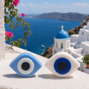 May include: Two eye-shaped candles. One is light blue with a white and black centre, the other is white with a blue and black centre. The candles are on a white surface with a scenic background of the sea and buildings.