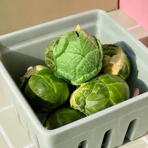 May include: A light green ceramic basket filled with several fresh Brussels sprouts and one green wax candle shaped like a Brussels sprout. The candle is lit with a small flame visible at the top, nestled among the real green vegetables.