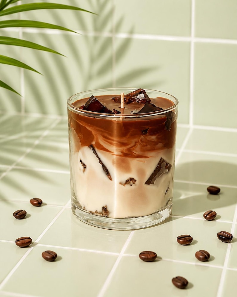 May include: A candle in a clear glass, designed to resemble an iced coffee drink. The candle has layers of white, brown, and dark brown wax, with wax cubes on top. Coffee beans are scattered around the base of the glass on a light green tiled surface.