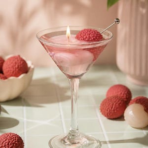 May include: A martini glass candle with a lit wick, surrounded by pink wax and a red lychee fruit. A small silver cocktail pick holds another lychee. Several whole lychees and a bowl of lychees are in the background.