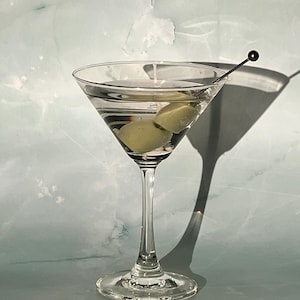 May include: A classic martini in a stemmed glass, featuring two green olives and a cocktail pick. The clear liquid is set against a light blue and white background, with a long shadow cast to the right.