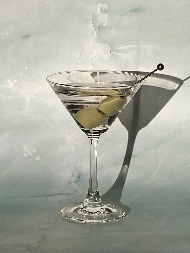 May include: A classic martini in a stemmed glass, featuring two green olives and a cocktail pick. The clear liquid is set against a light blue and white background, with a long shadow cast to the right.