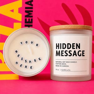 May include: Two candles on a pink background. One candle is in a white container with the words "YOUR MESSAGE HERE" spelled out in small black letter beads. The other candle is in a glass jar with a wooden lid and a label that reads "HIDDEN MESSAGE".
