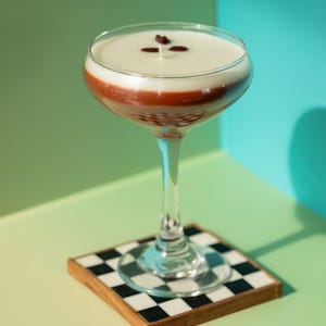 K&ouml;nnte beinhalten: Ein geschichteter Cocktail in einem Stielglas, mit einer cremigen, wei&szlig;en Oberseite, einer rotbraunen Mittelschicht und einer klaren Basis. Garniert mit drei Kaffeebohnen. Das Glas steht auf einem quadratischen Untersetzer mit schwarz-wei&szlig;em Schachbrettmuster.