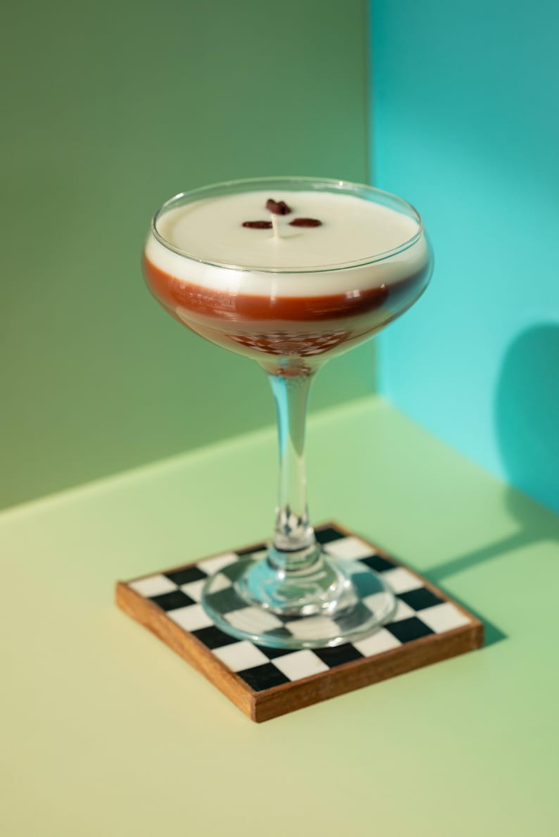 K&ouml;nnte beinhalten: Ein geschichteter Cocktail in einem Stielglas, mit einer cremigen, wei&szlig;en Oberseite, einer rotbraunen Mittelschicht und einer klaren Basis. Garniert mit drei Kaffeebohnen. Das Glas steht auf einem quadratischen Untersetzer mit schwarz-wei&szlig;em Schachbrettmuster.