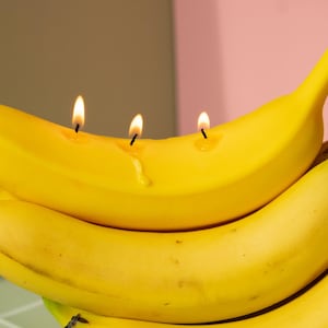 以下が含まれることがあります： 熟した黄色のバナナに3本の火の灯ったキャンドルが埋め込まれた、ユニークなキャンドルアレンジメント。バナナは他のバナナの束の上に置かれ、遊び心があり、予想外の装飾品となっています。