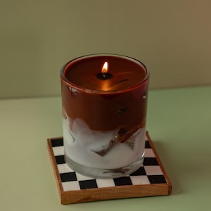May include: A lit candle in a clear glass with a two-tone appearance, featuring a dark brown top and a white, marbled bottom. The candle sits on a square coaster with a black and white checkered pattern.