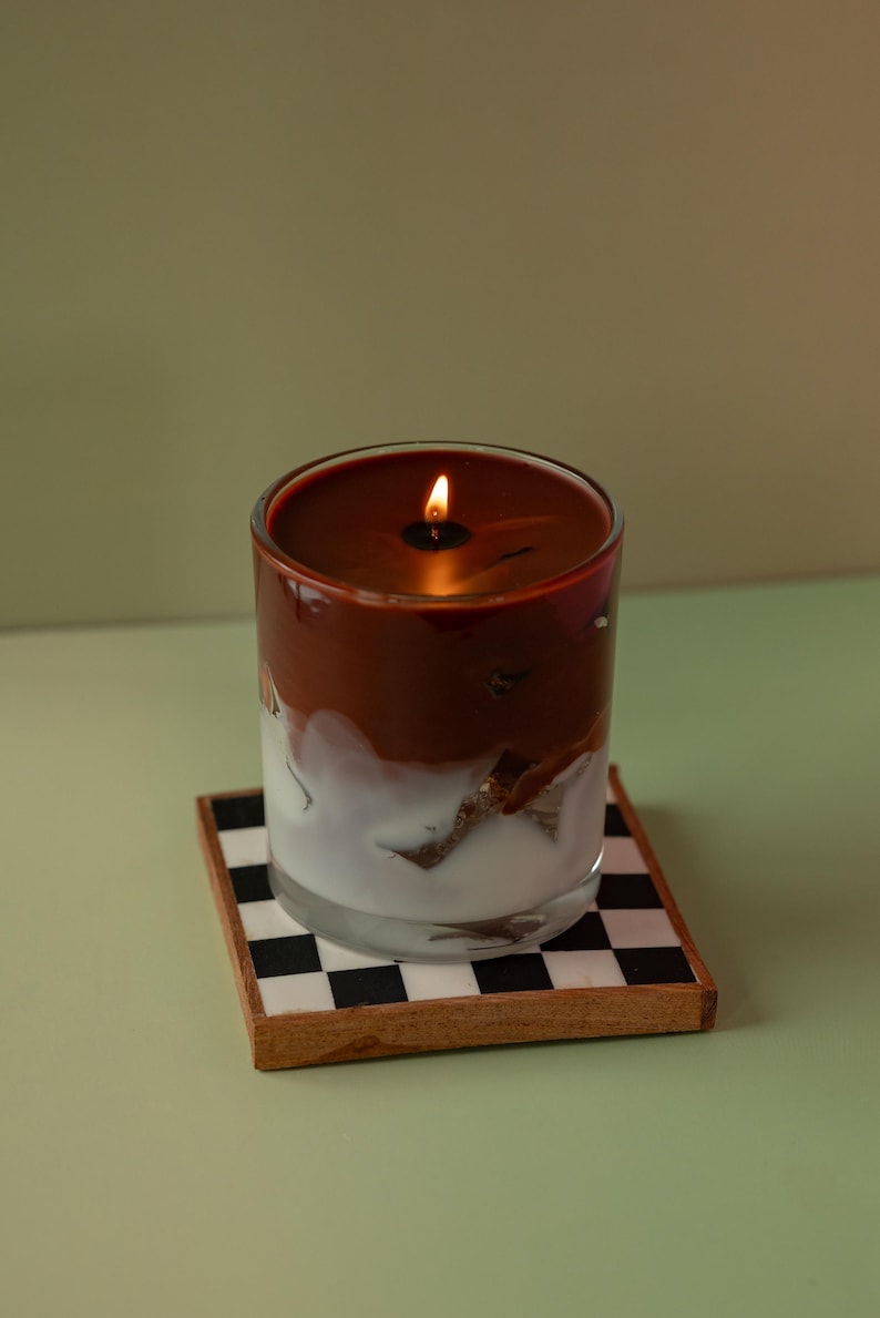 May include: A lit candle in a clear glass with a two-tone appearance, featuring a dark brown top and a white, marbled bottom. The candle sits on a square coaster with a black and white checkered pattern.