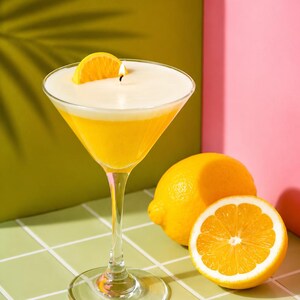 May include: A martini glass candle with a yellow wax base and a white top, topped with a lemon slice and a lit wick. Two whole lemons and a lemon half are next to the glass. The background is green and pink.