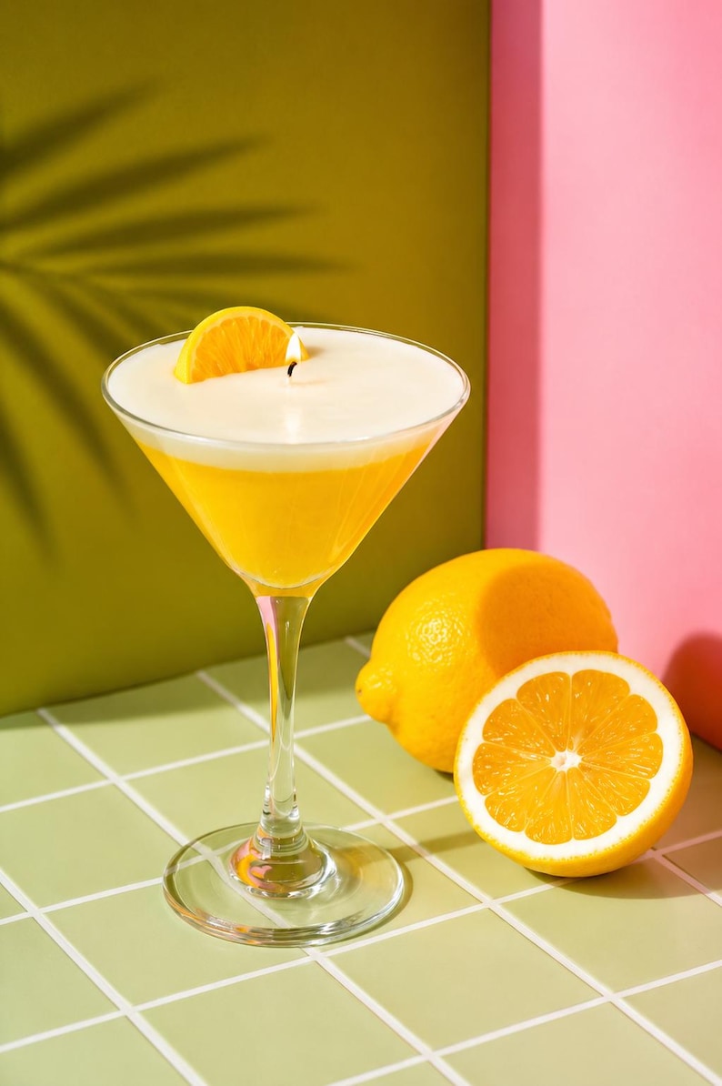 May include: A martini glass candle with a yellow wax base and a white top, topped with a lemon slice and a lit wick. Two whole lemons and a lemon half are next to the glass. The background is green and pink.