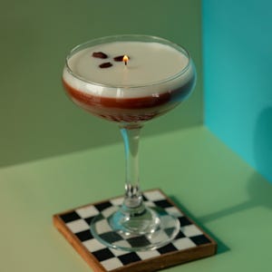 K&ouml;nnte beinhalten: Eine Cocktailglas-Kerze mit einer wei&szlig;en oberen Schicht, einer roten Mittelschicht und einem klaren Stiel. Die Kerze hat drei Kaffeebohnen obenauf und einen brennenden Docht. Sie steht auf einem quadratischen Untersetzer mit schwarz-wei&szlig;em Schachbrettmuster.