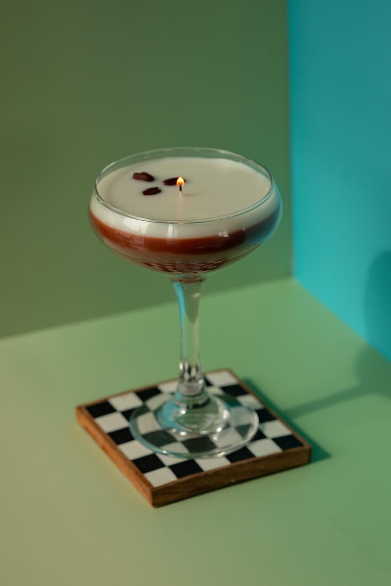 K&ouml;nnte beinhalten: Eine Cocktailglas-Kerze mit einer wei&szlig;en oberen Schicht, einer roten Mittelschicht und einem klaren Stiel. Die Kerze hat drei Kaffeebohnen obenauf und einen brennenden Docht. Sie steht auf einem quadratischen Untersetzer mit schwarz-wei&szlig;em Schachbrettmuster.