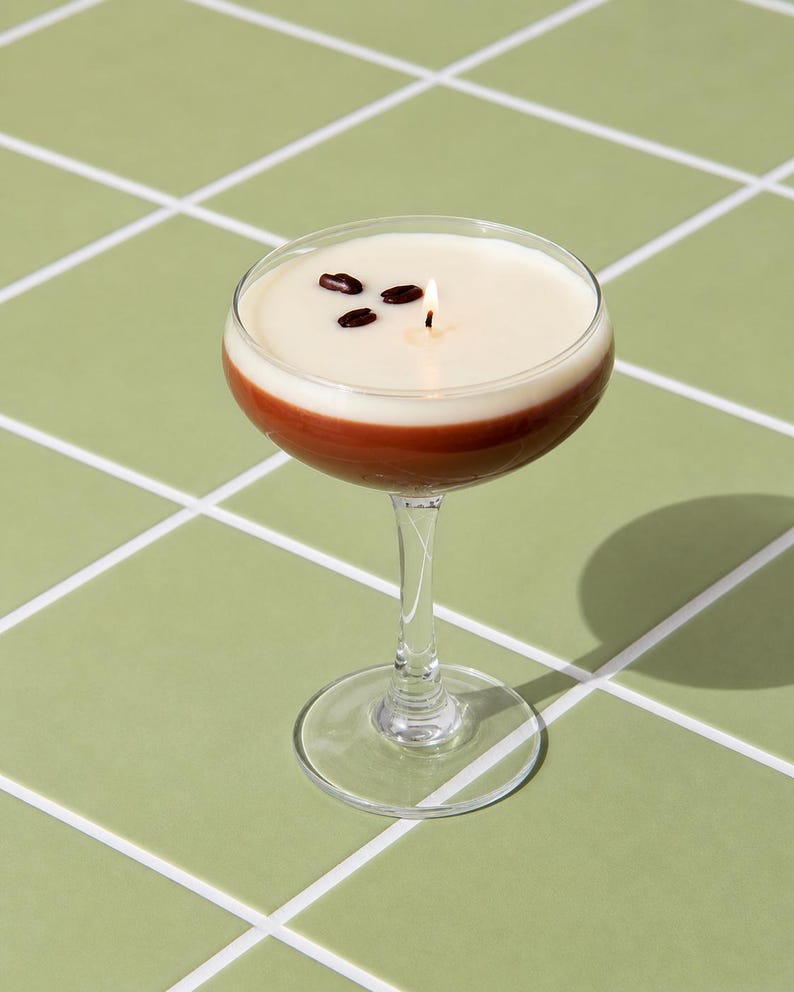 K&ouml;nnte beinhalten: Eine kerzenf&ouml;rmige Cocktailkerze mit braunem Boden, cremewei&szlig;er Oberseite und drei Kaffeebohnen. Ein angez&uuml;ndeter Docht ist sichtbar. Die Kerze steht auf einem klaren Glasstiel und -fu&szlig; vor einem gr&uuml;nen Fliesenboden.