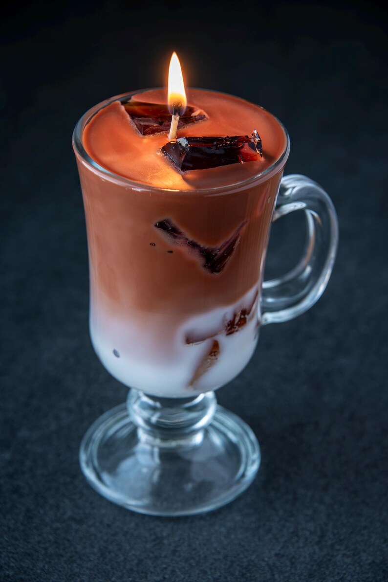 May include: A glass mug filled with a layered chocolate and white drink. The drink is topped with chocolate squares and a lit candle.