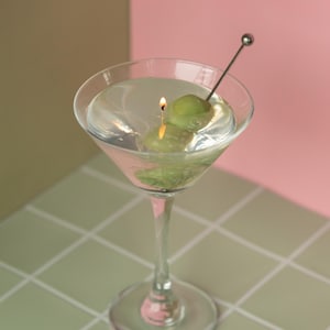 May include: A clear martini glass with a lit candle and a green olive. The candle is placed inside the glass, which is filled with a clear liquid. A silver cocktail pick holds the olive. The background is a mix of pink and brown.