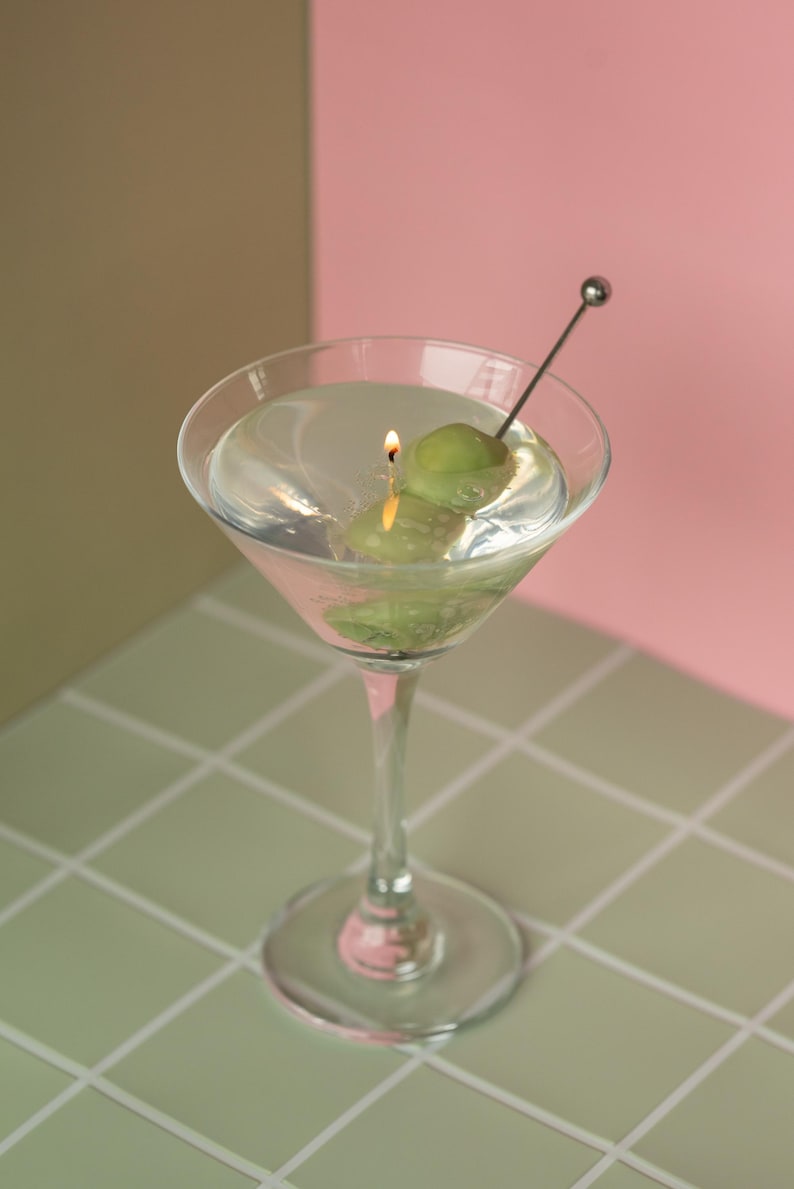 May include: A clear martini glass with a lit candle and a green olive. The candle is placed inside the glass, which is filled with a clear liquid. A silver cocktail pick holds the olive. The background is a mix of pink and brown.