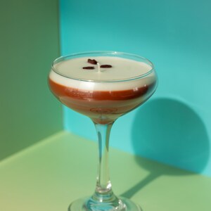 K&ouml;nnte beinhalten: Ein klares Cocktailglas gef&uuml;llt mit einem geschichteten Getr&auml;nk. Die untere Schicht ist dunkelbraun, bedeckt mit einer wei&szlig;en Schicht und garniert mit drei dunkelbraunen Kaffeebohnen. Das Glas steht auf einer gr&uuml;nen und blauen Oberfl&auml;che.